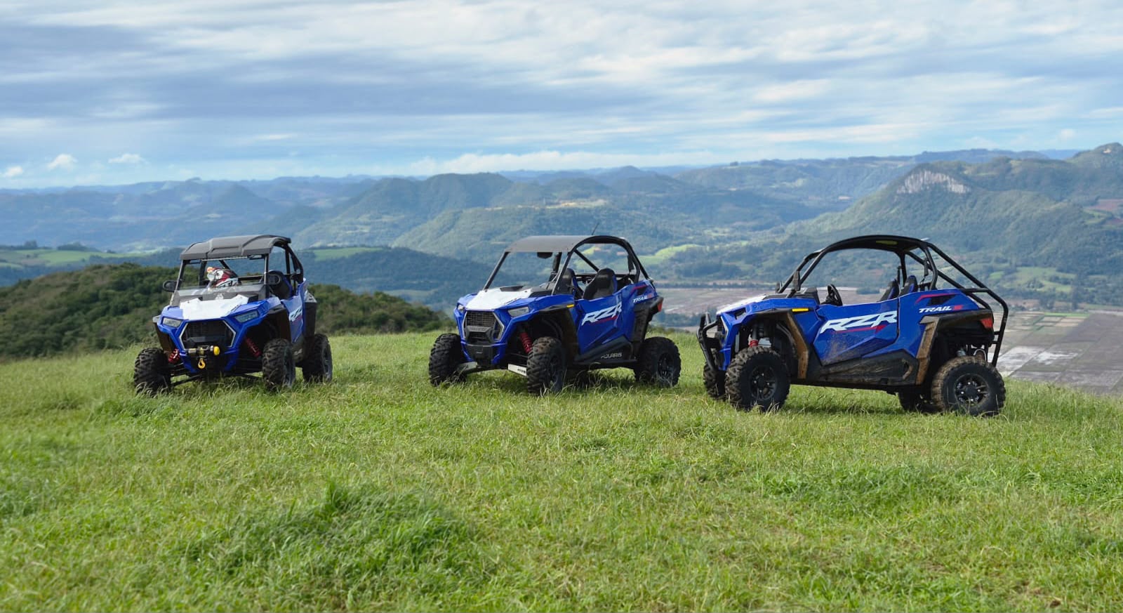 UTVs azuis alinhados na grama, com céu, mata e serra ao fundo