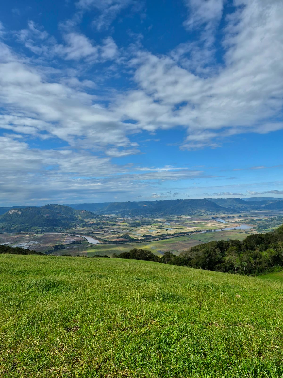 Vista vertical do vale da Quarta Colônia sob um céu azul com nuvens, com campo verde em primeiro plano