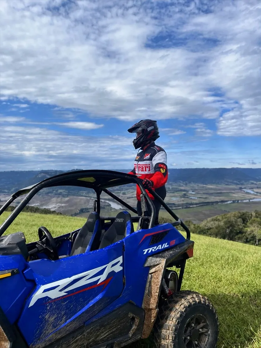 Pessoa de capacete em pé ao lado de um Polaris RZR azul, no alto de um campo com o vale ao fundo